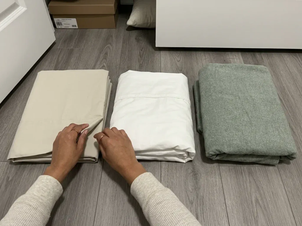 Three folded cotton sets on a closet floor, one crisp, one slumped, one napped flannel