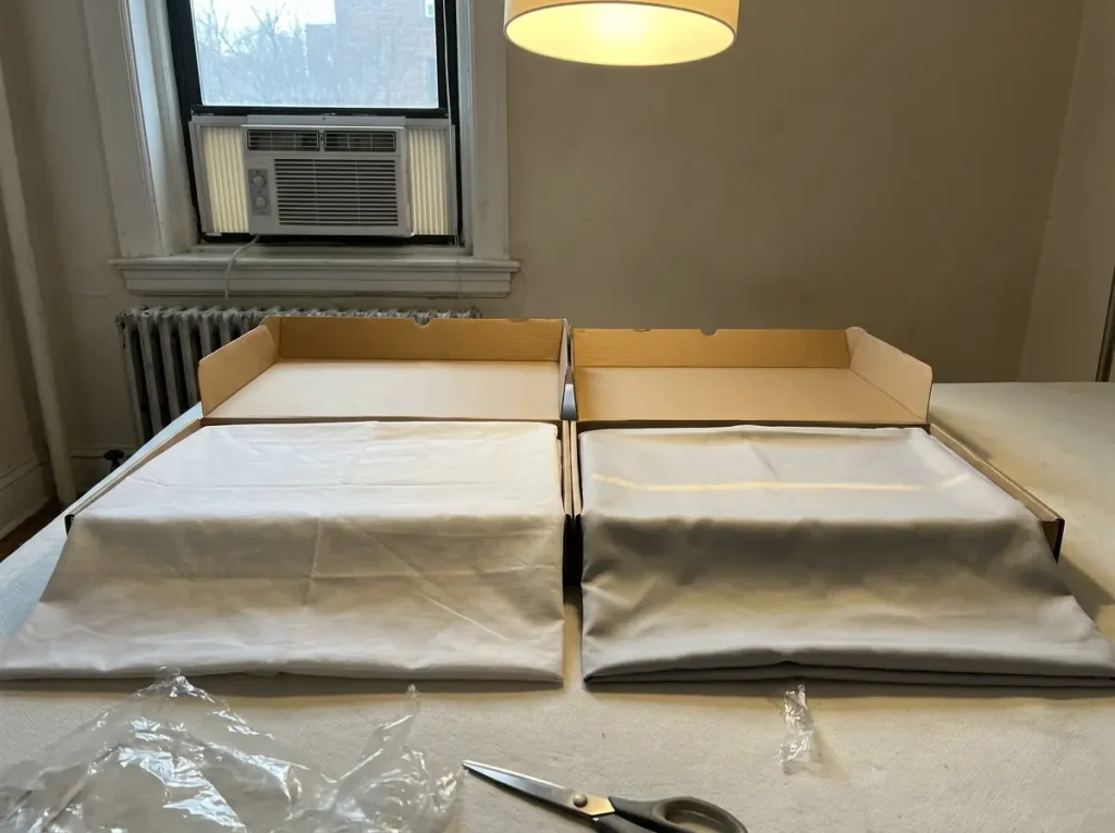 Two sheet packages side by side on a bed, one crisp cotton and one slick synthetic