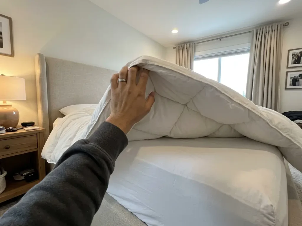 A hand lifting the edge of a lofted comforter so the thickness of the fill shows against a bright window