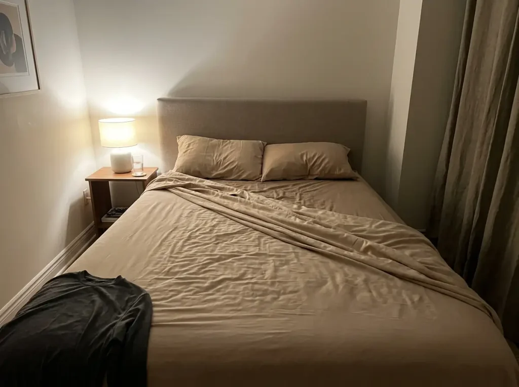 Microfiber top sheet pushed back at three in the morning, lamp lit beside a studio bed