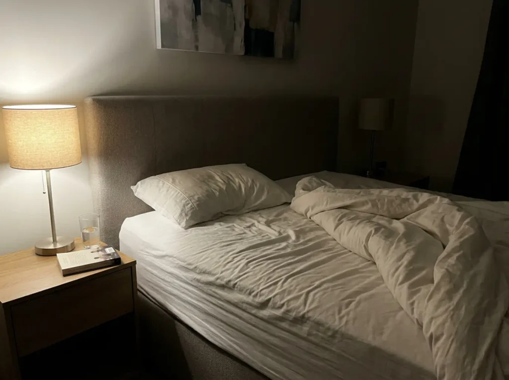 Rumpled sheet on one side of a bed at night, lit by a single bedside lamp