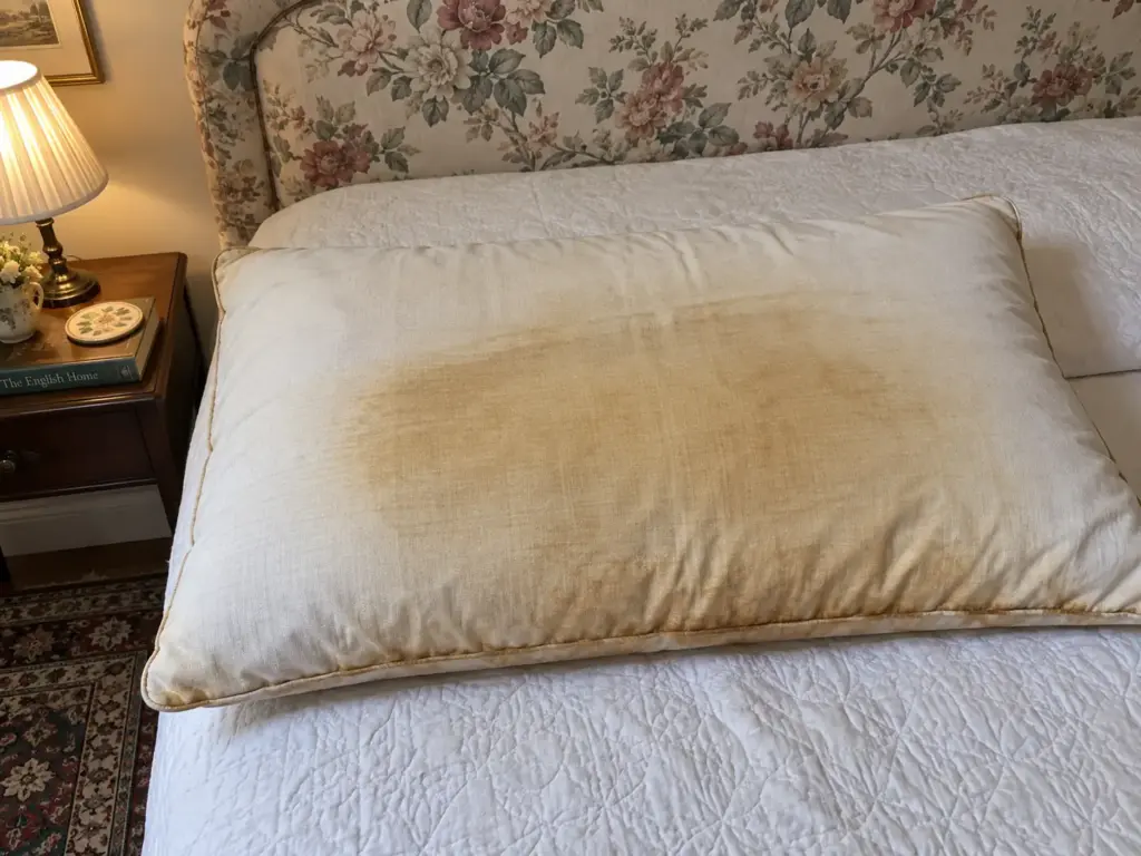 Close view of a bare cotton pillow showing yellowing deepest along the seam and where the head rests