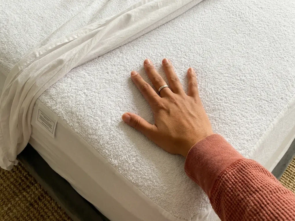 A hand pressed flat on the cotton terry face of a mattress protector with the fitted sheet turned back