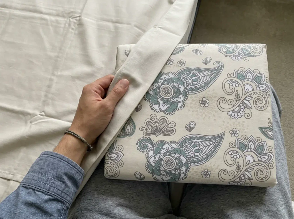 Hands holding a percale sheet set half out of its packaging on a guest bed, the open weave visible in flat daylight
