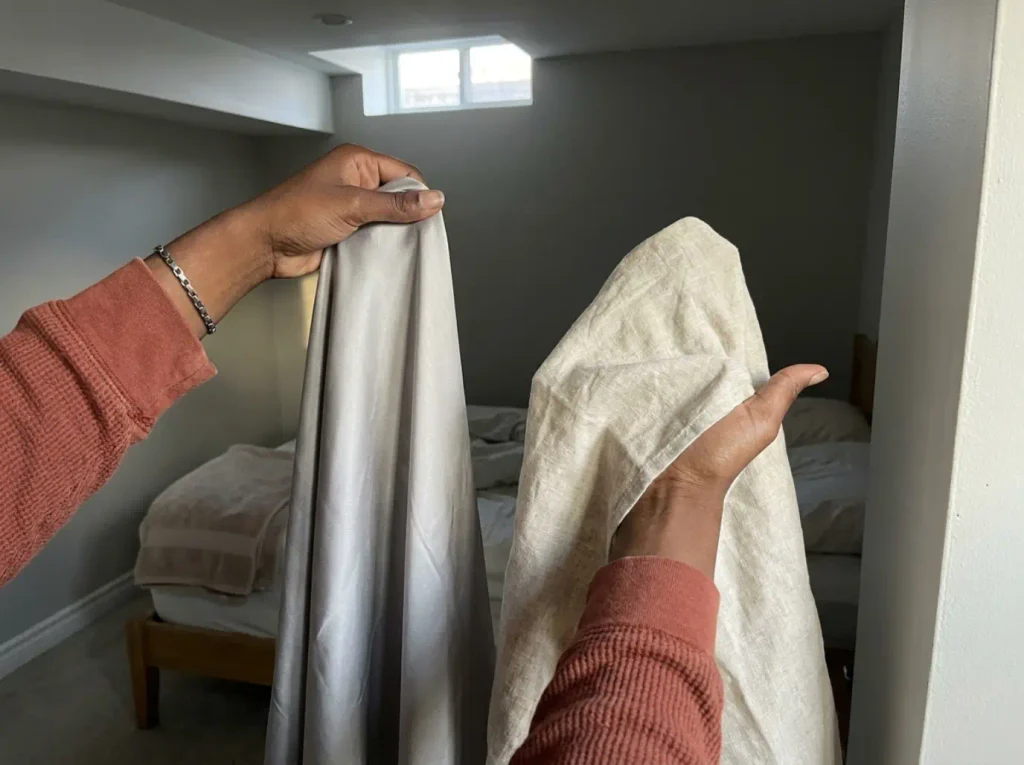 Two hands holding a corner of bamboo sheet and a corner of linen sheet side by side to compare