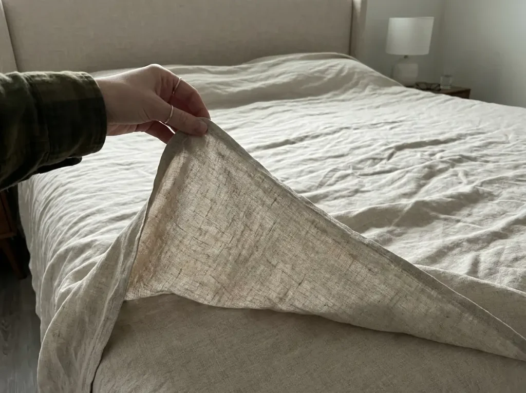 A hand pinching a corner of washed linen sheeting to show its weight and open weave