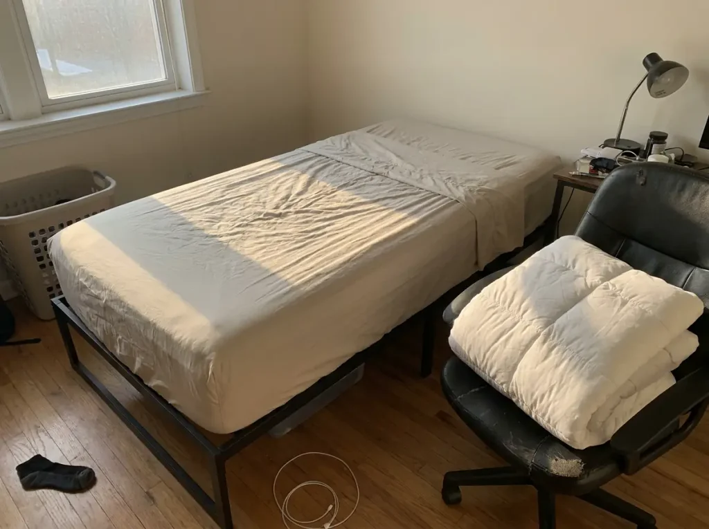 A bed made with only a fitted and top sheet, the comforter folded on a chair beside it