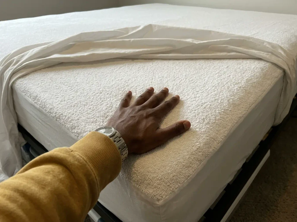 A hand flat on a cotton terry mattress protector with the fitted sheet peeled back off the corner of a bed
