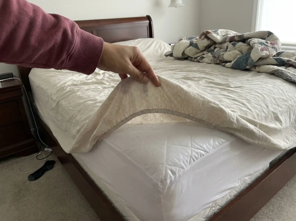 A fitted sheet peeled back at the mattress corner showing the quilted protector underneath