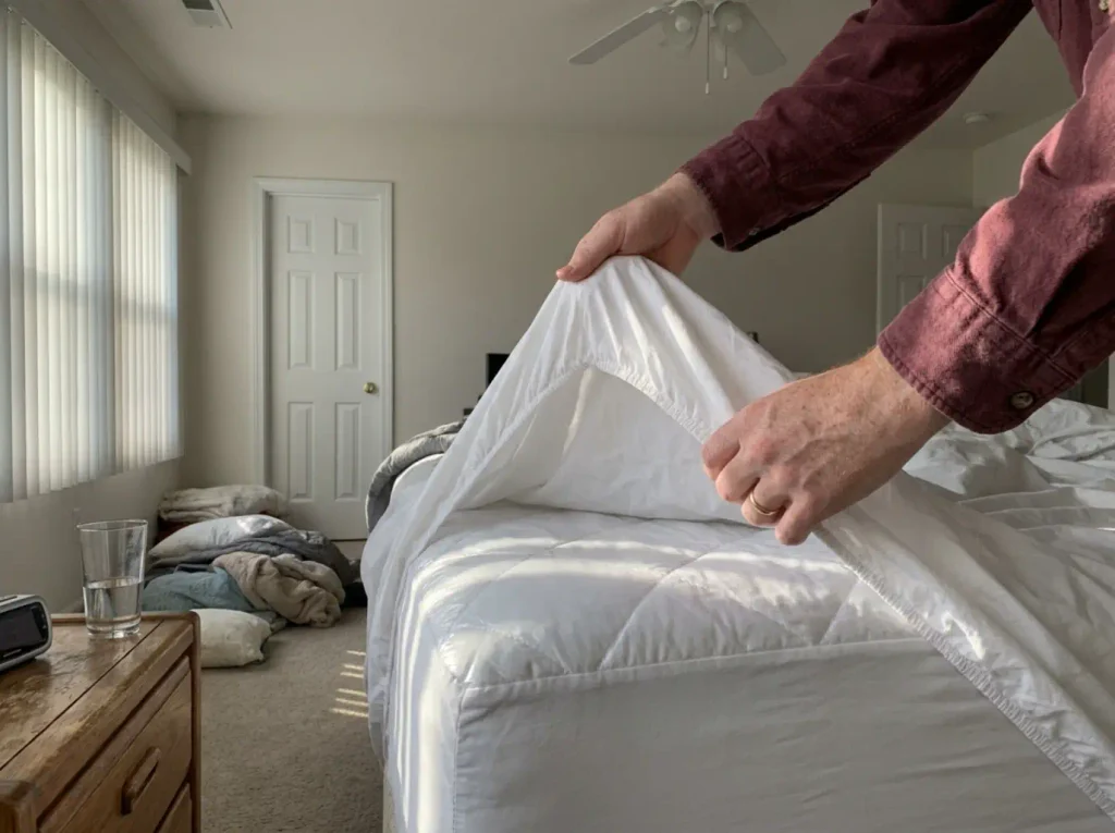 A fitted sheet peeled back off the mattress corner to show the protector underneath