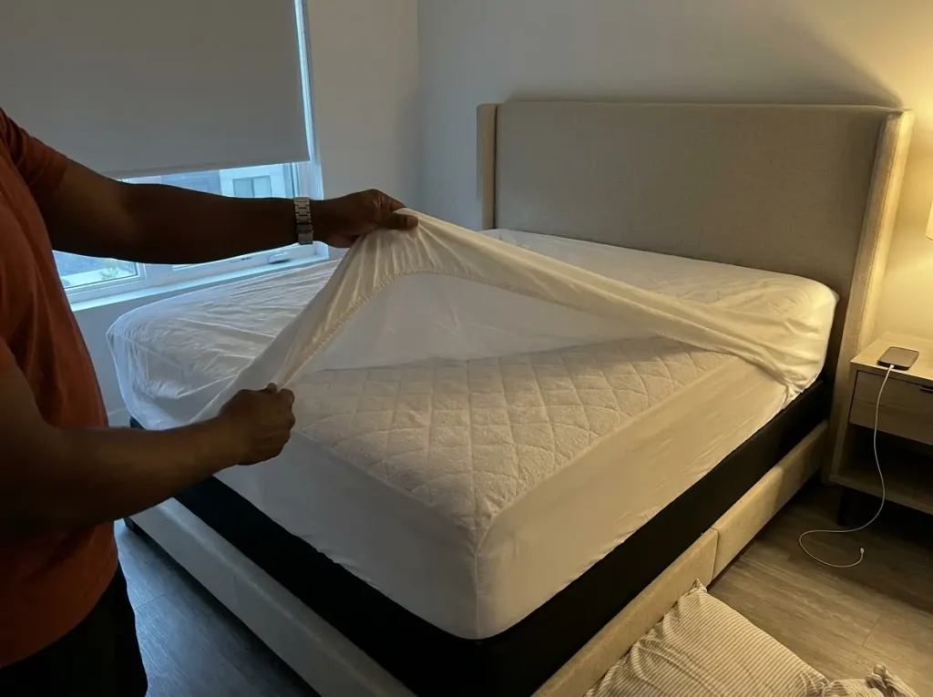 A fitted sheet peeled back at the corner to reveal the mattress protector underneath the bedding