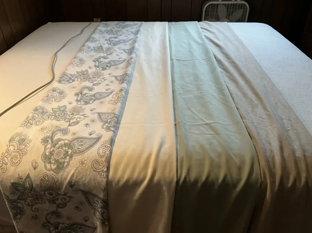 Four sheet fabrics laid overlapping on a stripped mattress under one raking lamp to compare surfaces